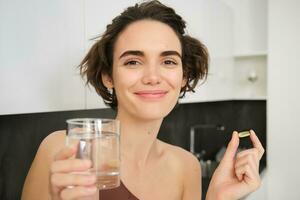 dieta suplementos e saudável estilo de vida. jovem mulher levando Vitamina c, d ômega-3 com vidro do água, em pé dentro roupas esportivas, bebendo depois de exercite-se Treinamento dentro dela cozinha foto