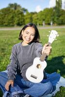 música e instrumentos. sorridente ásia menina mostra dela branco cavaquinho, senta dentro parque e tocam pequeno guitarra foto