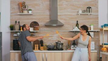 casal tendo Diversão dentro a cozinha esgrima com grande colheres durante café da manhã vestindo pijamas. alegre despreocupado alegre engraçado amantes, brigando de madeira colher, vínculo jogos luta esgrima, feliz estilo de vida foto