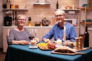 Senior casal olhando às Câmera durante relação celebração com festivo jantar dentro cozinha. aniversário, lazer, aposentado, família, romântico foto