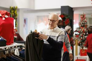 Mais velho cliente olhando às tecido dentro roupas loja, verificação material antes comprando roupas em desconto. pessoa visitando compras Centro durante feriado temporada, Veja para Natal presentes. foto