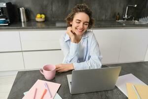 jovem mulher trabalhador autonomo trabalho a partir de lar, parece às computador portátil tela e sorrisos, faz dever de casa dentro cozinha foto