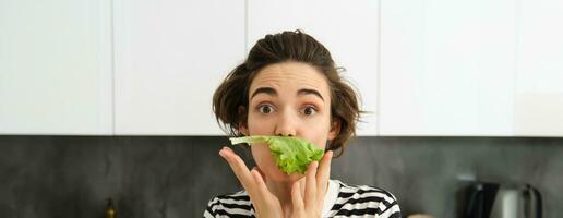 fechar acima do engraçado fofa mulher, vegetariano comendo alface folha e sorridente, conceito do saudável dieta, menina curtidas vegetais, em pé dentro a cozinha foto