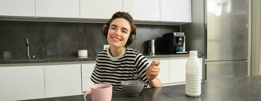 retrato do feliz sorridente mulher comendo cereais com leite, bebendo chá e ouvindo música dentro sem fio fones de ouvido, goza dela manhã, tendo café da manhã sozinho dentro a cozinha foto