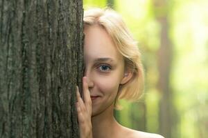 lindo menina espreitar Fora a partir de atrás uma árvore dentro a floresta foto