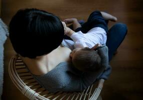 jovem mulher amamentação dela pequeno filho enquanto sentado dentro uma acolhedor poltrona foto