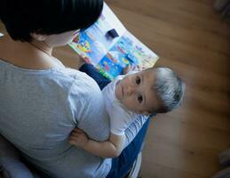 jovem mãe com pequeno filho lendo livro enquanto sentado dentro uma cadeira dentro a quarto foto