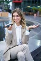 sorridente o negócio mulher registro voz mensagem, Falando para dentro microfone em Móvel telefone, sentado perto fonte em rua foto