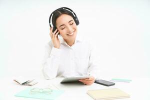 pessoas e ambiente de trabalho. sorridente ásia mulher senta dentro escritório, aluna ouvindo música dentro fones de ouvido e segurando digital tábua, branco fundo foto