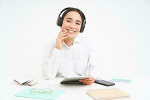 pessoas e ambiente de trabalho. sorridente ásia mulher senta dentro escritório, aluna ouvindo música dentro fones de ouvido e segurando digital tábua, branco fundo foto