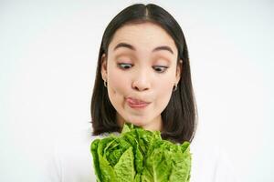 saudável Comida e vegano estilo de vida. fechar acima retrato do sorridente ásia mulher, parece feliz, mostra dela face com alface, come repolho, branco fundo foto