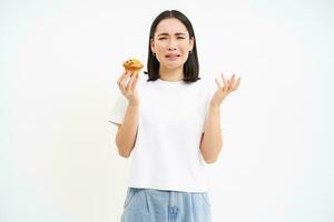 retrato do infeliz coreano mulher parece às Bolinho com triste face, menina em dieta hipócrita comer pastelaria, conceito do saudável comida, branco fundo foto