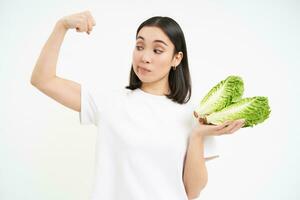 dieta e saúde. sorridente ásia mulher mostra dela bíceps, Forte corpo e repolho dentro mãos, come legumes para saudável nutrição, branco fundo foto