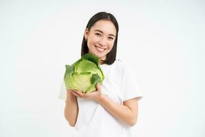 cintura acima retrato do sorridente ásia mulher, segurando repolho, recomenda comendo vegetais, saudável orgânico verde comida, isolado em branco fundo foto