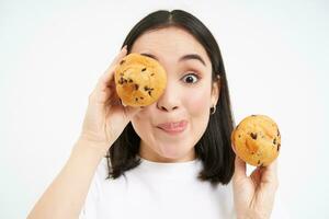 fechar acima do fofa ásia menina com Bolinho, mostra bolos perto face e parece tentado para mordida sobremesa, isolado em branco fundo foto