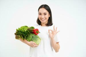 retrato do sorridente ásia mulher, segurando grupo do legumes e faz OK sinal, recomenda verde nutritivo Comida Entrega aplicativo, branco fundo foto