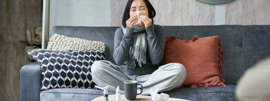 jovem ásia mulher ficando às lar, sentindo-me indisposto, pegando uma frio, doente deixar, espirros, bebendo quente chá e levando medicação foto