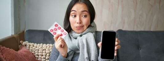 feliz ásia mulher pegando frio, mostrando Smartphone tela e pílulas, recomendando conectados médico aplicativo, gp ou médico clínica em telefone, segurando drogas, ficando às casa com gripe foto