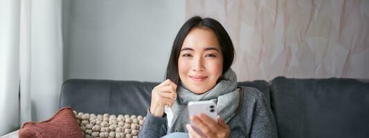 retrato do coreano mulher sente doente, segurando Smartphone, chamando médico gp para pegue prescrição, apanhado frio, ficando às lar, usando Móvel telefone foto