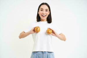 feliz jovem coreano mulher, mostrando dois bolos de copo sobre peito, sorridente e olhando feliz, pastelaria e padaria conceito foto