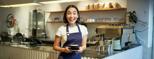 retrato do sorridente ásia fêmea barista, fazer café, segurando copo do chá e levando isto para cafeteria cliente, vestindo avental, em pé perto contador foto