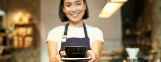 sorridente ásia menina barista, em pé dentro avental uniforme, dando você copo do café, fez uma beber para cliente dentro cafeteria foto