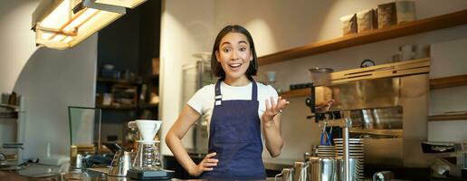 retrato do menina barista dentro avental, parece surpreso, pontos com mão e olhares fixos espantado às Câmera, trabalho atrás contador e cervejas café foto