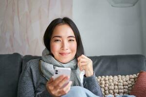 retrato do coreano mulher sente doente, segurando Smartphone, chamando médico gp para pegue prescrição, apanhado frio, ficando às lar, usando Móvel telefone foto