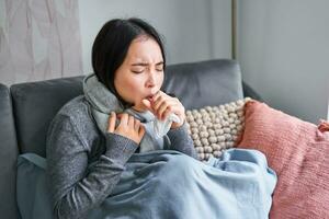 ásia mulher tosse, sentindo-me doente, sentado debaixo cobertor com scard dentro frio apartamento, salvando em aquecimento isto inverno, pegando gripe foto