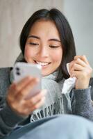 retrato do coreano mulher sente doente, segurando Smartphone, chamando médico gp para pegue prescrição, apanhado frio, ficando às lar, usando Móvel telefone foto