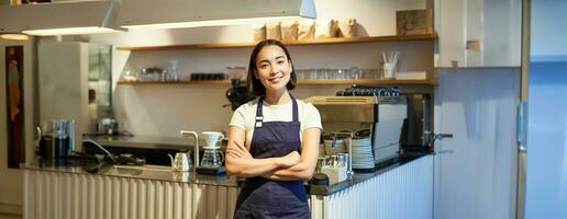 sorridente ásia menina barista, em pé perto contador dentro cafeteria, vestindo companhia avental, Cruz mãos e parece confiante foto