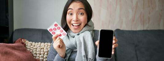 conectados médicos e assistência médica. sorridente coreano mulher mostra Móvel telefone tela e medicamento, pegando frio, ficando às casa e recomendando Smartphone gp foto