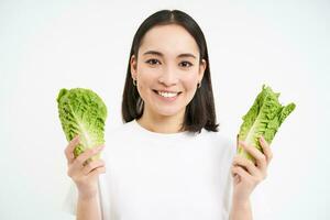 feliz coreano mulher vegetariano, sorridente e mostrando repolho, comendo alface em dieta, conduz ativo estilo de vida, branco fundo foto