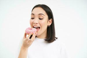 fechar acima do sorridente satisfeito ásia mulher, come envidraçado Rosa rosquinha, goza comendo saboroso donut, branco estúdio fundo foto