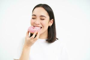 fechar acima do sorridente satisfeito ásia mulher, come envidraçado Rosa rosquinha, goza comendo saboroso donut, branco estúdio fundo foto