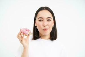 fechar acima do sorridente satisfeito ásia mulher, come envidraçado Rosa rosquinha, goza comendo saboroso donut, branco estúdio fundo foto