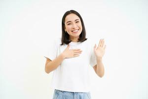 amigável, sorridente coreano mulher, mostra 1 mão acima e coloca Palma em coração, faz promessa, introduzir ela mesma, branco fundo foto