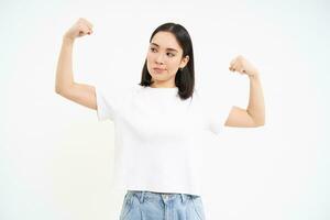 sorridente ásia mulher, flexão bíceps, olhando atrevido e forte, mostrando dela força músculos, branco estúdio fundo foto