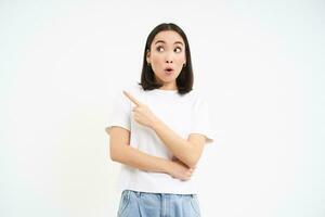 surpreso coreano garota, pontos esquerda e parece com espantado face expressão, carrinhos dentro camiseta isolado em branco fundo foto