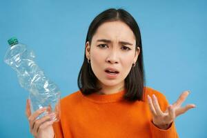 retrato do confuso mulher, segurando e mostrando plástico garrafa, sem noção porque pessoas estão não reciclando, azul fundo foto