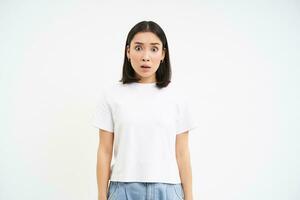 imagem do preocupado japonês mulher, suspiros e olhares fixos com choque às Câmera, poses dentro branco camiseta contra estúdio fundo foto