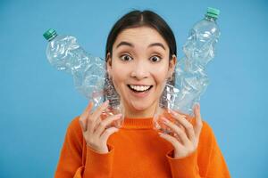 entusiasmado sorridente mulher mostra dois reciclável garrafas, reciclando plástico e olhando feliz, azul fundo foto