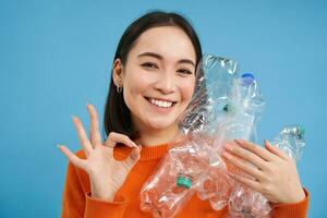 reciclando é ótimo. sorridente jovem mulher, segurando garrafas, mostra OK sinal, aprova eco verde estilo de vida, Ordenação lixo, azul fundo foto