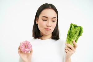 saudável estilo de vida conceito. jovem mulher decidir entre saudável vegetais, repolho e delicioso envidraçado rosquinhas, branco fundo foto