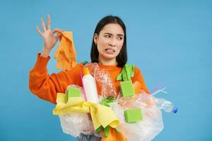 coreano mulher parece às fedido látex luva, detém plástico lixo, limpa casa e reciclar, azul fundo foto
