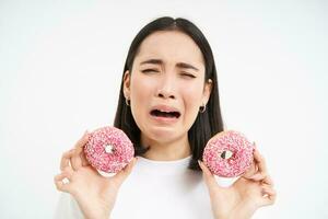 chateado soluçando menina hipócrita comer lixo comida, mostra delicioso envidraçado rosquinha, quer para morder, branco fundo foto