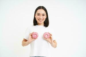 fofa sorridente mulher detém dois donuts perto dela peito e olhando feliz, comendo sobremesa, branco estúdio fundo foto