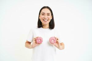 engraçado coreano mulher posando com dois delicioso envidraçado rosquinhas em peito, rindo e sorridente, come lixo comida, saboroso sobremesa foto