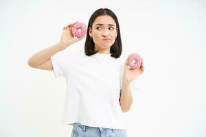 imagem do ásia menina detém dois rosquinhas, pensando do quebra dela dieta, parece com dúvida às donuts, branco fundo foto