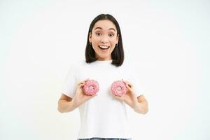 engraçado coreano mulher posando com dois delicioso envidraçado rosquinhas em peito, rindo e sorridente, come lixo comida, saboroso sobremesa foto